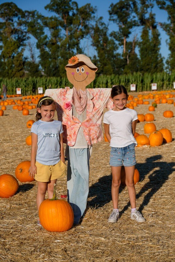 Two kids posing with a smiling scarecrow at a pumpkin patch in fall